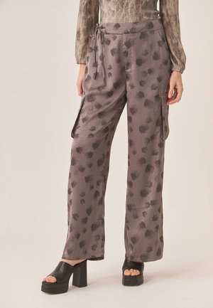 Mujer vistiendo pantalones cargo holgados de color gris con estampado, con bolsillos laterales y sandalias negras de plataforma con tacón alto, de pie contra un fondo liso.