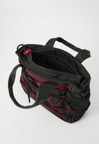 Sac en toile noir avec des coutures rouges, poignée supérieure, fermeture éclair et plusieurs compartiments. Corps texturé avec des détails en plis.