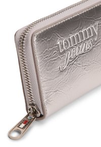 Ασημένιο μεταλλικό πορτοφόλι με υφή στην επιφάνεια, ανάγλυφο λογότυπο "tommy jeans" και μπρούτζινο φερμουάρ με έμβλημα στην τσιμπίδα.
