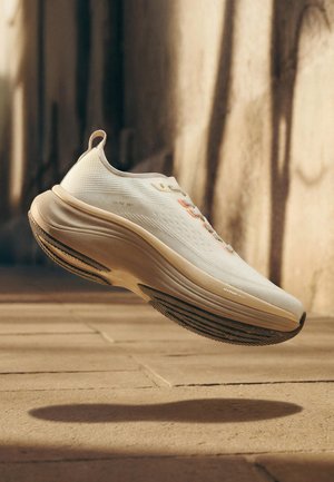 Světle béžová sportovní teniska s tlustou podešví označenou "HYPERTECH", zavěšená nad dlaždicovou podlahou a vrhající stín.