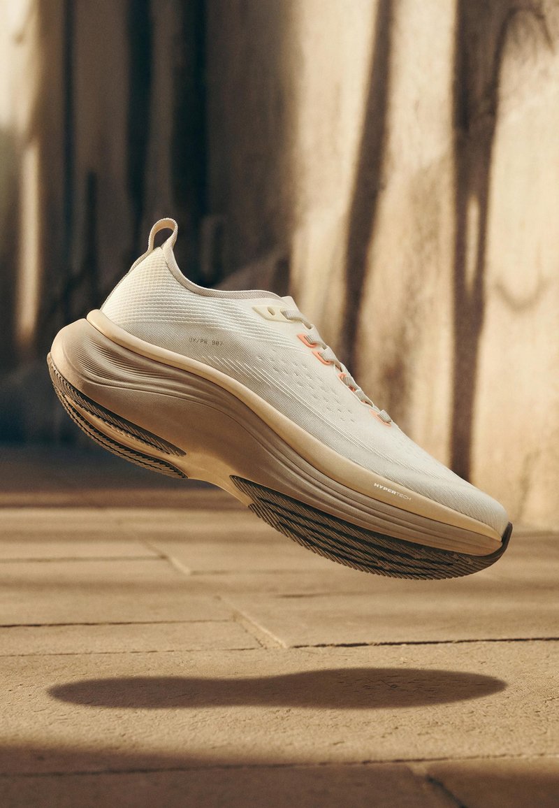 Baskets sportives beige clair avec une semelle épaisse marquée "HYPERTECH", suspendues au-dessus d'un sol carrelé projetant une ombre.