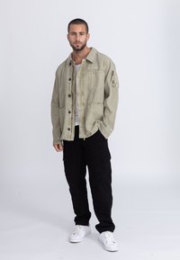 Hellgrüne Denimjacke mit Fronttaschen, Knopfverschluss und lockerer Passform, kombiniert mit schwarzen Cargo-Hosen und weißen Sneakers.