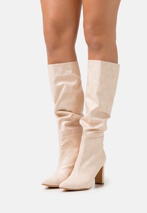 Jambes portant des bottes en cuir beige jusqu'aux genoux, souples, à bouts pointus et talons blocs, se tenant sur un fond blanc.