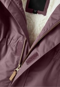 Fialová zipová bunda s měkkou bílou fleecovou podšívkou, zlatými detaily na zipu a látkovými nášivkami blízko zipu pro bezpečné uzavření.