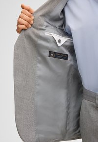 Una persona tiene aperto un giacca da completo grigia, mostrando la fodera liscia grigia con tasca interna ed etichetta del marchio con la scritta "Total Travel".