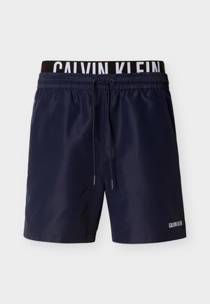 Tumesinised ujumistrikoodid elastse vöökohaga, millel on valge logo, paelaga kinnis ja küljetaskud. Sile tekstuur, pikkus üle põlve.