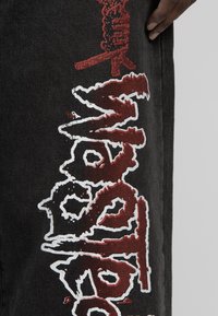 Svarta denimbyxor med broderad röd och vit grafisk text. Sömmen visar en texturerad finish som tillför visuell intressivität.