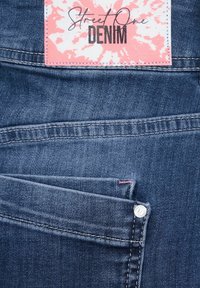 Jeans i denim med en mörkblå färg, som har en framficka, kontrasterande sömmar och en rosa och vit etikett som står "Street One DENIM."