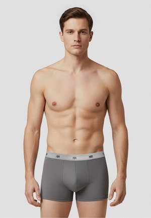 Skjorteløs mand iført tætsiddende grå boksertrusser med en lys grå taljebånd med gentagende logo, stående mod en ensfarvet hvid baggrund.