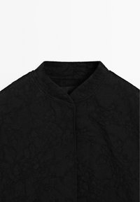 Svart texturerad tygjacka med subtilt blommönster och mandarin-krage, visad från halsringningen till mitten av bröstet.