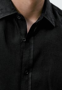 Zbliżenie na szyję i górną część klatki piersiowej osoby w czarnej koszuli z guzikami i kołnierzykiem.