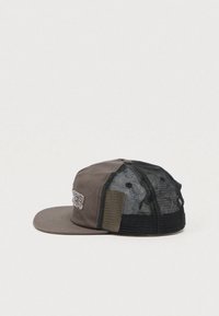 Καφέ και μαύρο πλεκτό καπέλο μπέιζμπολ με επίπεδη γείσο και κεντημένο έμβλημα στο μπροστινό μέρος, εμφανιζόμενο από το πλάι σε απλό φόντο.