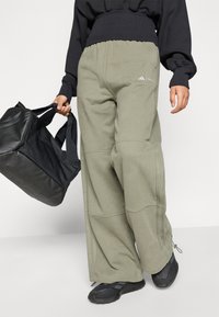 Personai, kurš valkā brīvus olīvu zaļas bikses ar logo, melnas kurpes, melnu garām piedurknēm topu, labajā rokā tur melnu sporta somu.