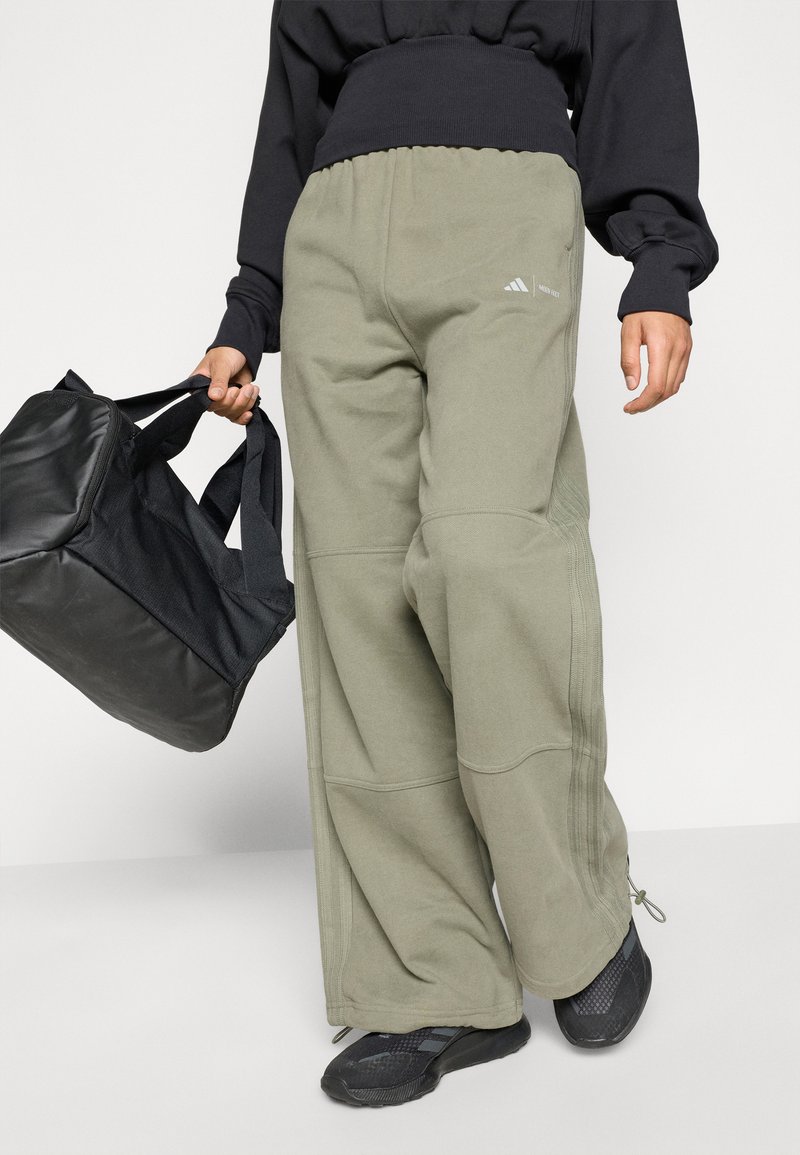 Persoon in losse olijfgroene broek met logo, zwarte schoenen, zwart langemouwen shirt, houdt een zwarte sporttas in de rechterhand.