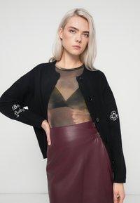 Melna pārāk liela izmēra kardigāna ar lielām pogām, ar izšūtu "Esi laimīgs" tekstu un dekoratīvu rakstu uz piedurknes, apvienots ar caurspīdīgu blūzi un bordo ādas svārkiem.