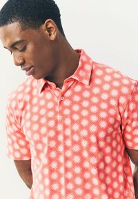 Camisa polo de manga corta en color coral, con un patrón de sol en rosa claro, cuello abotonado y textura de tela suave.