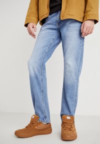 Vaqueros de denim azul claro con un corte de pierna recta, combinados con zapatillas de ante marrón. La tela tiene un acabado texturizado y un detalle mínimo.