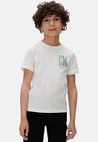 Camiseta de algodón blanca con un logo verde de CK, de manga corta y escote redondo. Combinada con pantalones negros. Textura suave y casual.