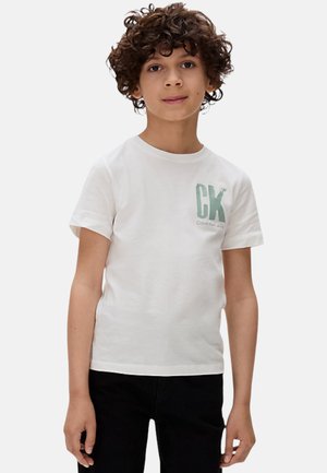 Camiseta de algodón blanca con un logo verde de CK, de manga corta y escote redondo. Combinada con pantalones negros. Textura suave y casual.