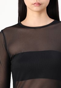 Haut long à manches transparentes noir avec un col rond et une bande noire unie sur la poitrine. Tissu léger et texturé.