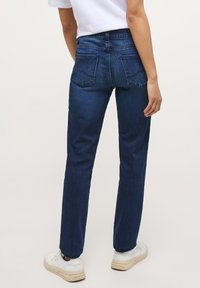 Osoba oblečená v tmavě modrých džínách a bílých teniskách, stojící zády k fotoaparátu na jednobarevném pozadí.