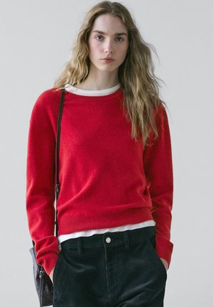 Ung kvinde med langt bølget hår iført en rød sweater over en hvid skjorte, sorte bukser og en skuldertaske mod en ensfarvet baggrund.