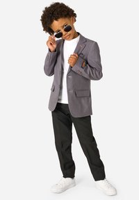 Chłopak z kręconymi włosami poprawia czarne okulary przeciwsłoneczne, ubrany w szary żakiet, białą koszulę, czarne spodnie i białe trampki, stojąc pewnie.