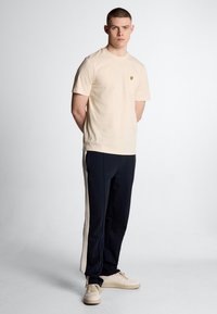 Beige katoenen t-shirt met een borstzak, gecombineerd met marineblauwe trainingsbroek met crèmekleurige zijkanten, en beige sneakers.