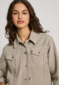 Giubbotto di jeans beige con chiusura a bottoni, colletto classico, due tasche sul petto e un tessuto leggermente strutturato. Design semplice con accenti minimi.