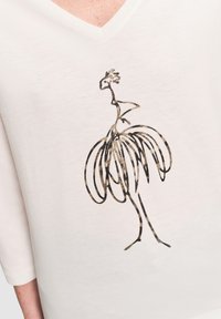 Maglietta bianca a V con un design astratto di uccello in nero e oro sul davanti. Il materiale appare morbido con una texture liscia.