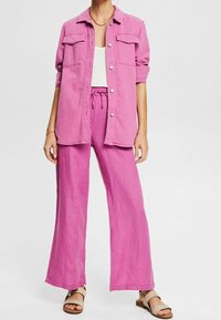 Femme portant une chemise rose ample à boutons avec manches retroussées, pantalon large rose assorti, débardeur blanc, collier chaîne et sandales beiges.