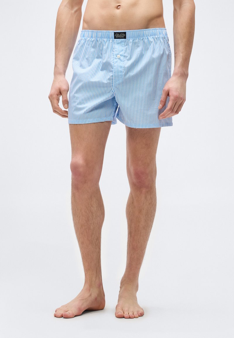 Άνδρας που στέκεται ξυπόλυτος φορώντας ελαφρώς  γαλάζια και λευκά κάθετα ριγέ μποξεράκια με κουμπί και ετικέτα στη μέση.