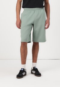 Μακριές πράσινες κοντές παντελόνες από βαμβάκι με ελαστική μέση, φτάνουν μέχρι τη μέση του μηρού, συνδυασμένες με μαύρα αθλητικά παπούτσια και λευκές κάλτσες μέχρι τον αστράγαλο.