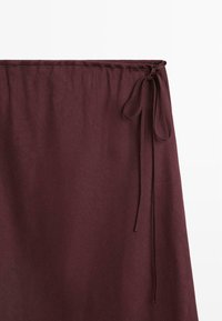 Mørk burgunder farvet stofnederdel med bindelukning i siden, glat tekstur og let rynkning ved taljen mod en ensartet hvid baggrund.