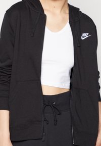 Svart zip-up hoodie med en åtsittande design, featuring en Nike-logotyp i vitt, dragsnöre i nederkant och mjukt, texturerat tyg.