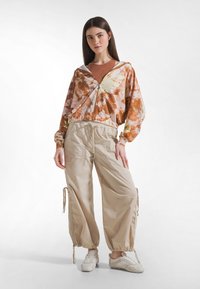 Farverig tie-dye lynlåstrøje med elastisk kant, kombineret med løstsiddende beige cargobukser med snore ved anklerne og sidelommer.