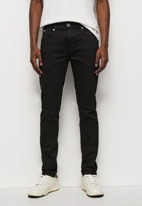 Crne slim-fit traperice od denima. Imaju kopčanje na dugme, pet džepova i jednostavan dizajn. Uparene su s bijelim tenisicama.