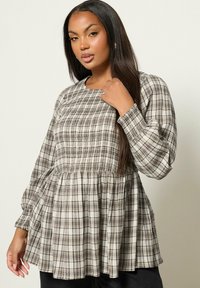 Vrouw met lang donker haar, gekleed in een beige en bruine geruite blouse met smock en lange pofmouwen tegen een effen achtergrond.