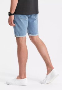 Pantalones cortos de mezclilla azul claro con dobladillos enrollados, que cuentan con dos bolsillos traseros y se combinan con zapatillas blancas sin cordones, mostrados de lado.
