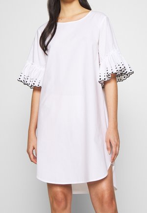Femme portant une robe blanche ample avec des manches volantées jusqu'au coude, décorées de pois noirs et de bords festonnés.