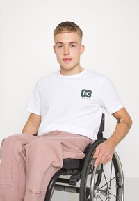 Camiseta de algodón blanca con estampado verde "03 TEAM," combinada con pantalones rosa claro, llevada por una persona en una silla de ruedas negra. Se aprecian texturas suaves.