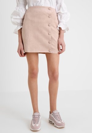 Femme portant une mini-jupe beige avec des boutons latéraux, un chemisier blanc à manches bouffantes et des baskets rose clair, debout devant un fond uni.