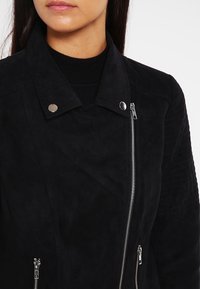 Veste en daim noir avec un col cranté, des accessoires en métal argenté, une fermeture zippée asymétrique et des textures sur les épaules.