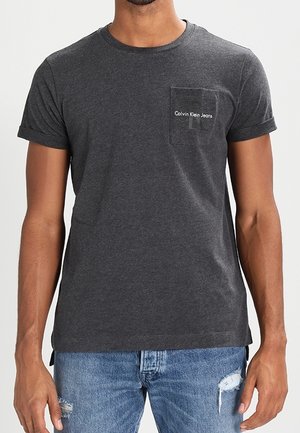 Homme portant un t-shirt gris foncé Calvin Klein Jeans avec poche et un jean bleu clair usé, face à l'avant avec les bras détendus.