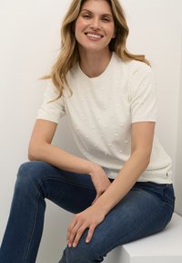 Strikket sweater i cremefarve med korte ærmer, der har opraised dot-mønstre. Bæres med mørkeblå jeans, siddende mod en hvid baggrund.