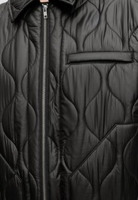 Gros plan d'une veste matelassée noire avec une couture ondulée, une fermeture éclair frontale et deux poches visibles, horizontale et inclinée.