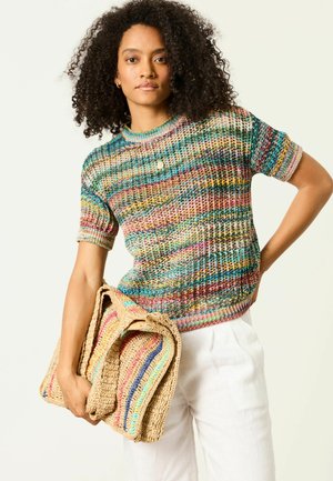 Woman with curly hair wears multicolored knit short-sleeve top and white pants, holding folded beige woven blanket with colorful stripes.