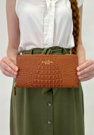 Brown crocodile-embossed leather wallet with a gold logo, zip closure, and a textured surface, held by a person in a white top and green skirt.