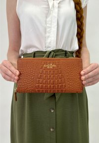 Pruun krokodillimustriga nahast rahakott, millel on kuldne logo, lukuga sulgumine ja tekstureeritud pind, mida hoiab inimene valges peatus ja rohelises seelikus.