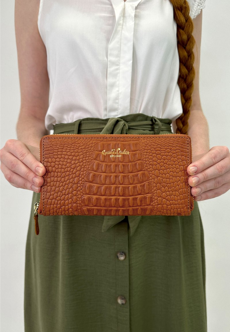 Pruun krokodillimustriga nahast rahakott, millel on kuldne logo, lukuga sulgumine ja tekstureeritud pind, mida hoiab inimene valges peatus ja rohelises seelikus.